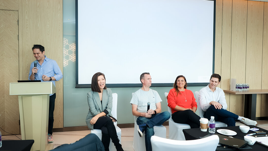 A man stands at a podium speaking into a microphone while four people, two women and two men, sit on chairs beside him, smiling and engaged, in front of a large blank screen in a conference room.