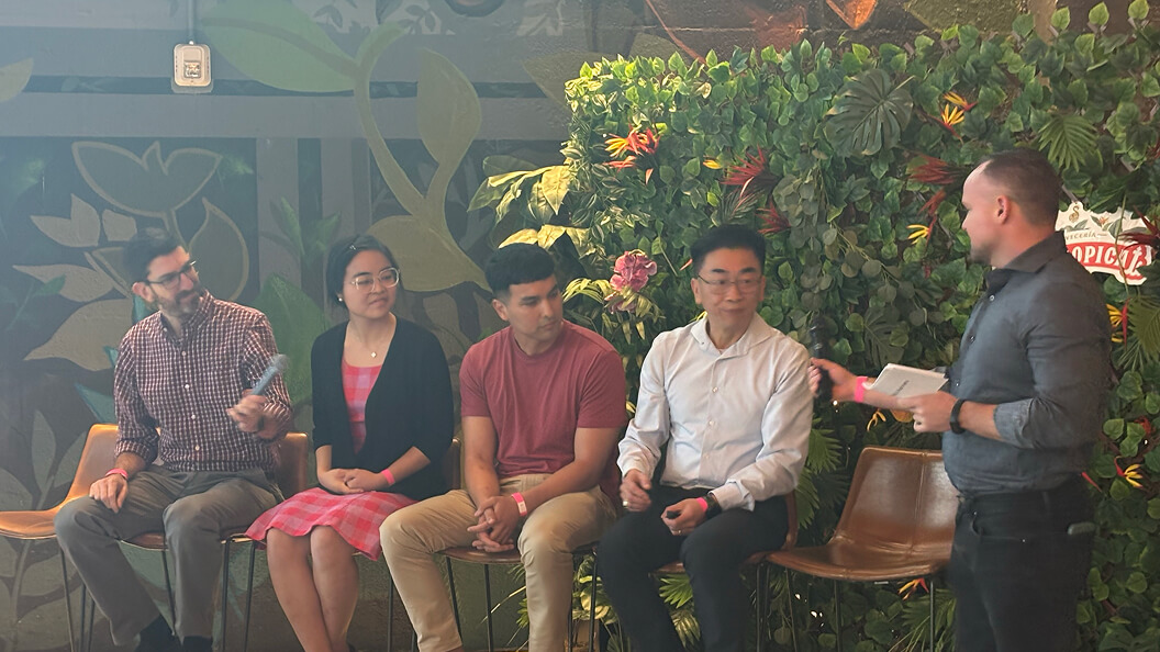 Four people are seated in a row on a stage with a leafy green backdrop, while a fifth person stands to the right, speaking and holding notes, engaging the seated group in conversation.