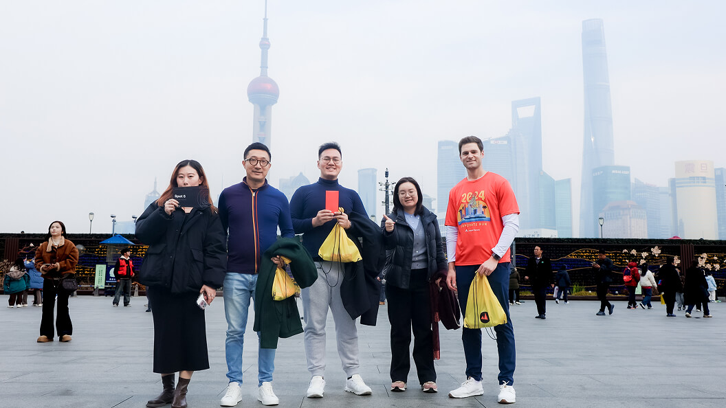 Five people stand posing outdoors in a city square with tall skyscrapers in the background on a cloudy day. They are holding yellow bags and smiling at the camera. Other people are visible further behind them.