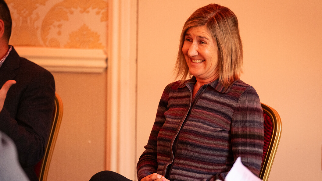 A woman with shoulder-length blonde hair sits on a chair, smiling, wearing a striped jacket. She is indoors, with ornate wallpaper and a cream-colored wall in the background.