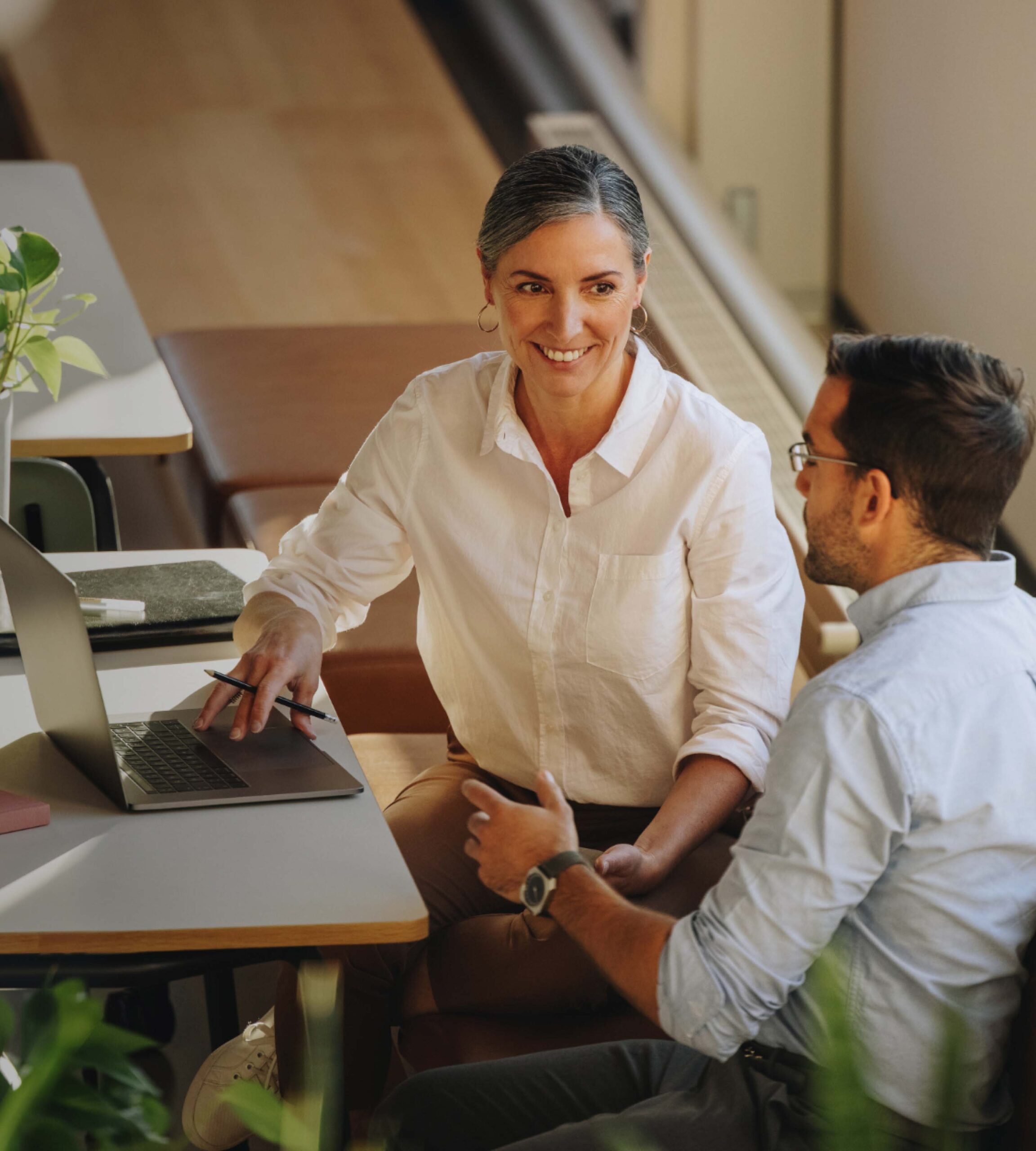 Two people sit at a table in a modern office, talking and smiling. One woman gestures at a laptop while the man listens attentively. Green plants and notebooks are visible on the table.