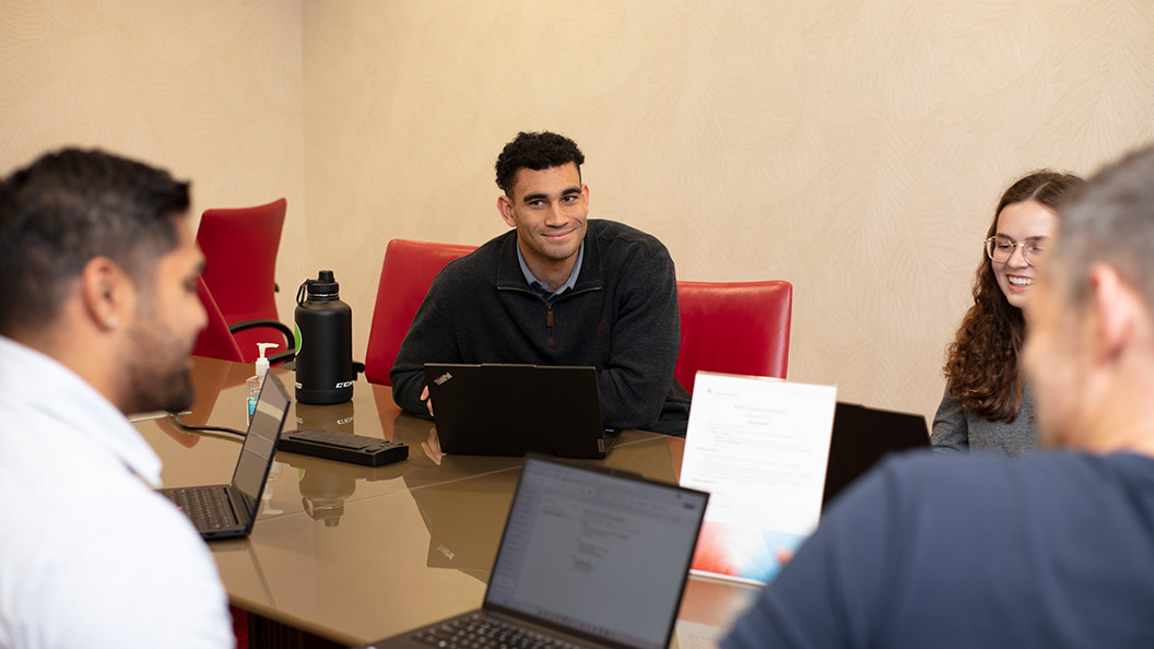 Four people sit around a conference table with laptops, engaged in discussion. One person is smiling at the center, while others look toward each other. A water bottle and paperwork are also on the table.