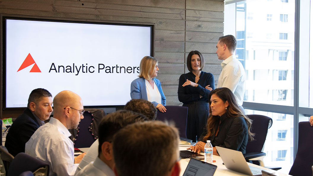 A group of professionals are gathered in a modern conference room. Some are seated around a table with laptops, while three people stand and talk in front of a screen displaying the Analytic Partners logo.