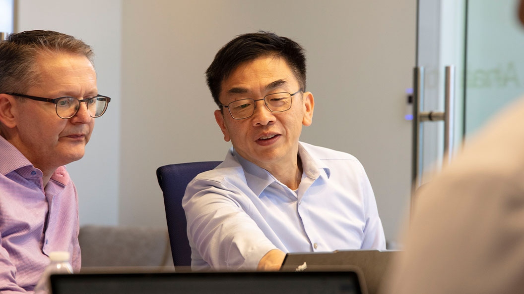 Two men in dress shirts sit at a table in an office, engaged in discussion. One points towards a laptop screen, while the other listens attentively. Another person is partially visible in the foreground.