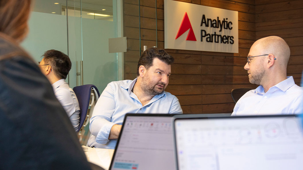 Two men in business attire have a conversation at a conference table in an office. A blurred laptop is in the foreground. A sign on the wood-paneled wall reads Analytic Partners.