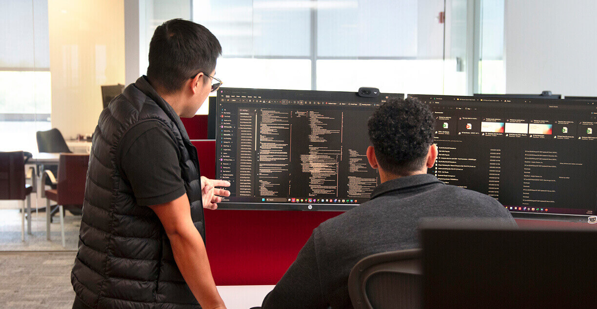 Two people sit at desks in an office, looking at large computer monitors filled with code and data. One person stands and points at the screen while the other is seated, both focused on the information displayed.
