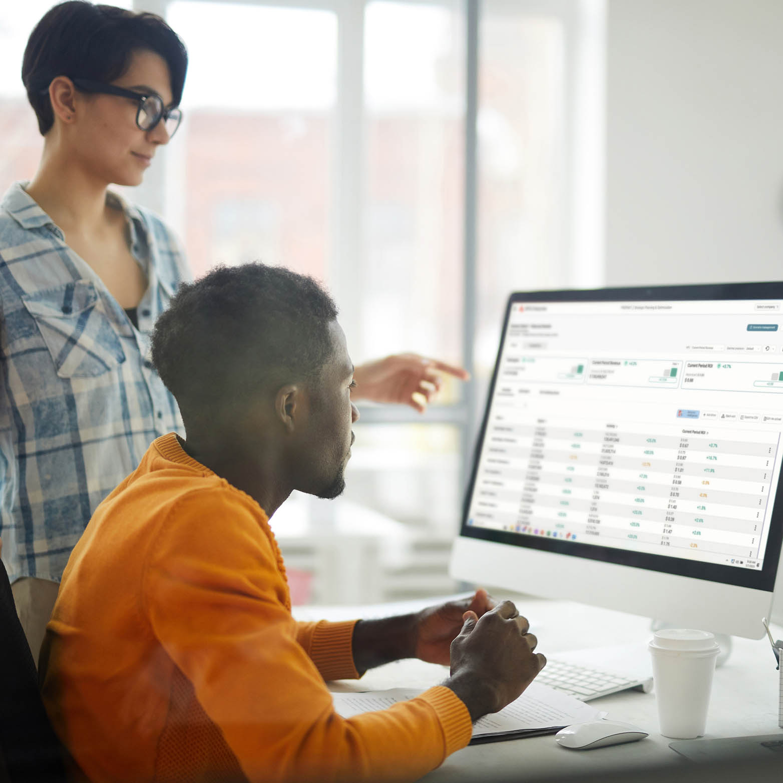 Two people work together at a desk, looking at a computer screen displaying data and charts. One sits and types while the other stands beside, pointing at the monitor. A coffee cup and notebook are on the desk.