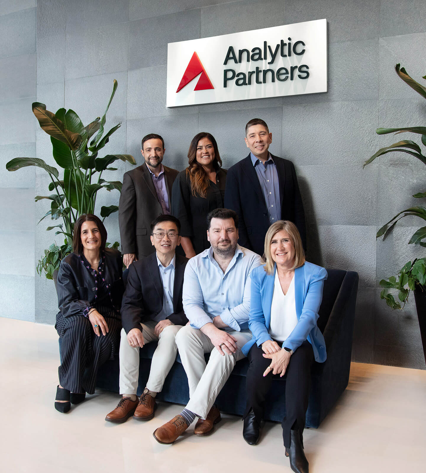 Seven professionally dressed people, three standing and four seated, pose in front of an Analytic Partners sign inside an office with green plants and light-colored flooring.